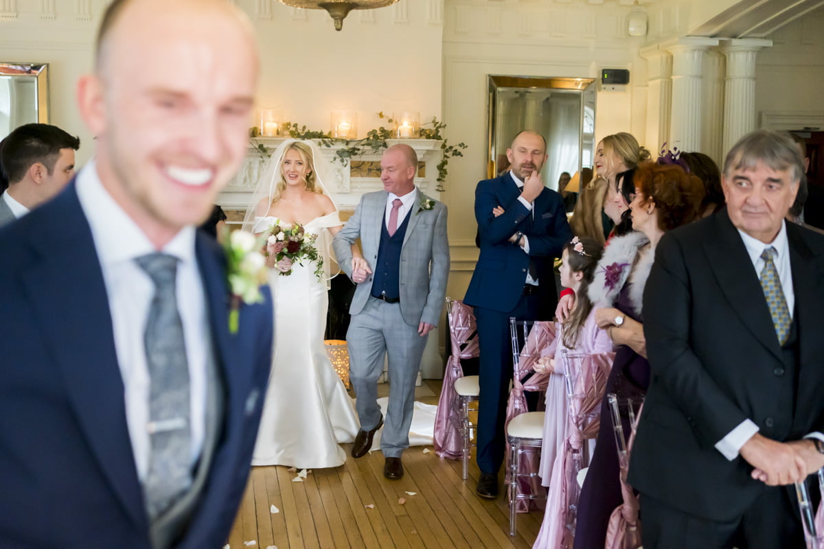The bride walks down the aisle with her father as the groom smiles