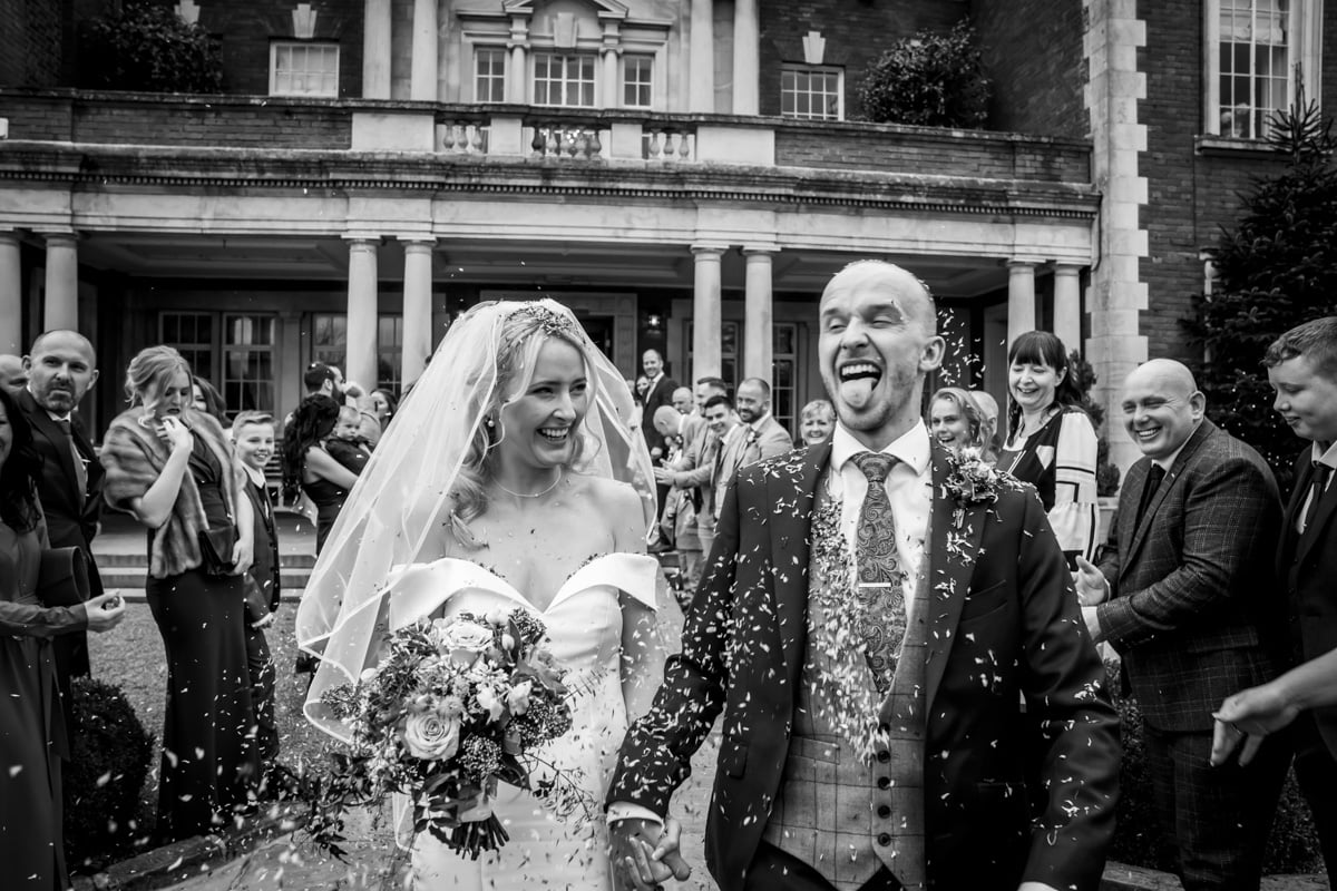 Guests shower the bride and groom as the groom sticks his tongue out with confetti at Eaves Hall