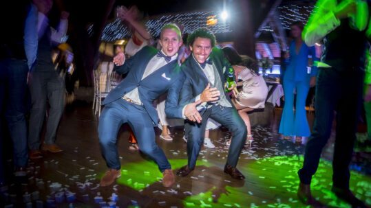 Two groomsmen dance together in the middle of the dancefloor, bathed in green light